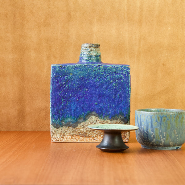 Mood image of a still life featuring three pieces of mid-20th century ceramics. The largest piece that stands centre and is square in shape is by Annikki Hovisaari. The small flying saucer shaped candlestick in the foreground is by Liisa Kaarina Hallamaa Larsen. Whilst the pot far right is a Danish design by Arne Bang. 