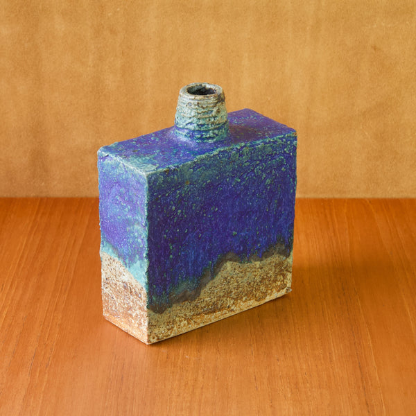 Top down image showing a large square shaped bottle vase stood on a wooden table. The blue glazed vase is by Finnish ceramicist Annikki Hovisaari who was born in 1918 in Tampere and died in 2004.