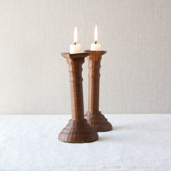 Two candlesticks woven in wicker and holding two lit candles. The form of the body is reminiscent of Doric columns found in Georgian architecture.